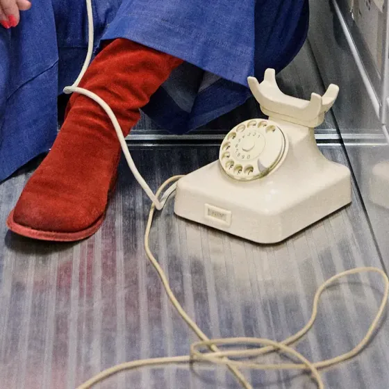 Ausschnitt: rote Stiefel, ein altes cremefarbenes Wählscheibentelefon auf dem Boden, das Kabel führt aus dem Bild.